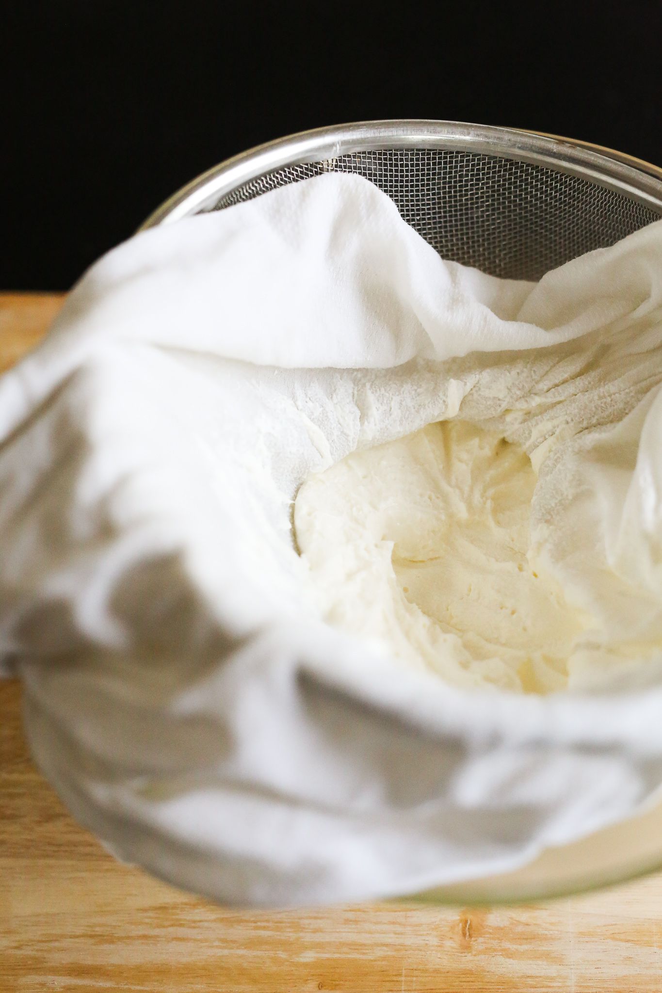 labneh in a white cloth-lined silver strainer sitting on cutting board on black table