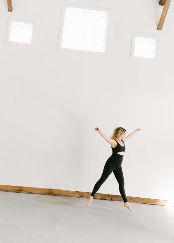 woman in unitard jumping in a loft space with three windows and concrete floors