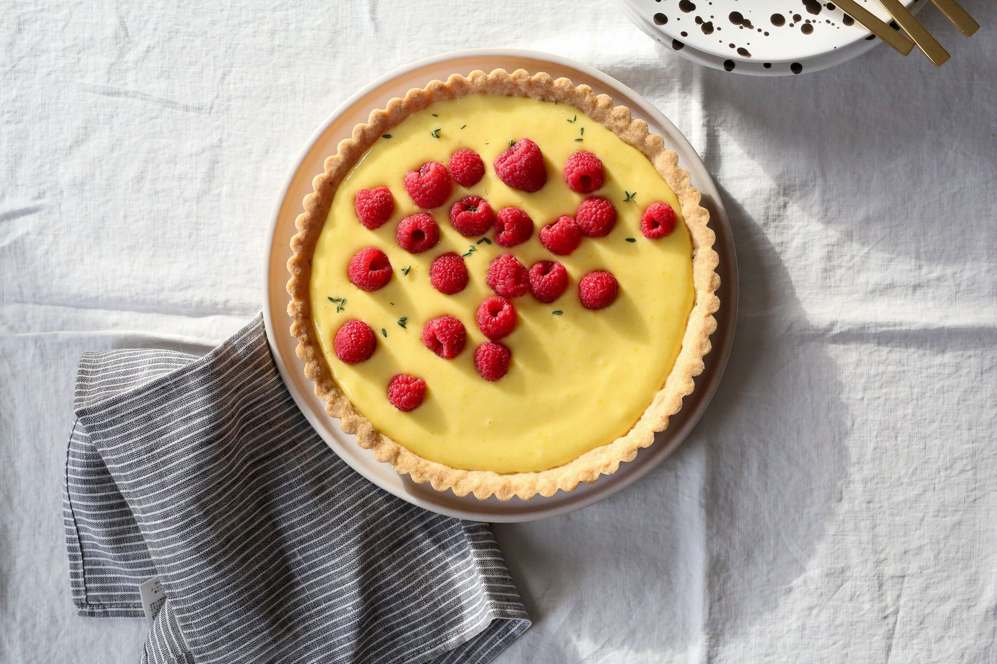 lemon tart with raspberries and thyme on white tablecloth with napkin and brown speckled plate gold forks