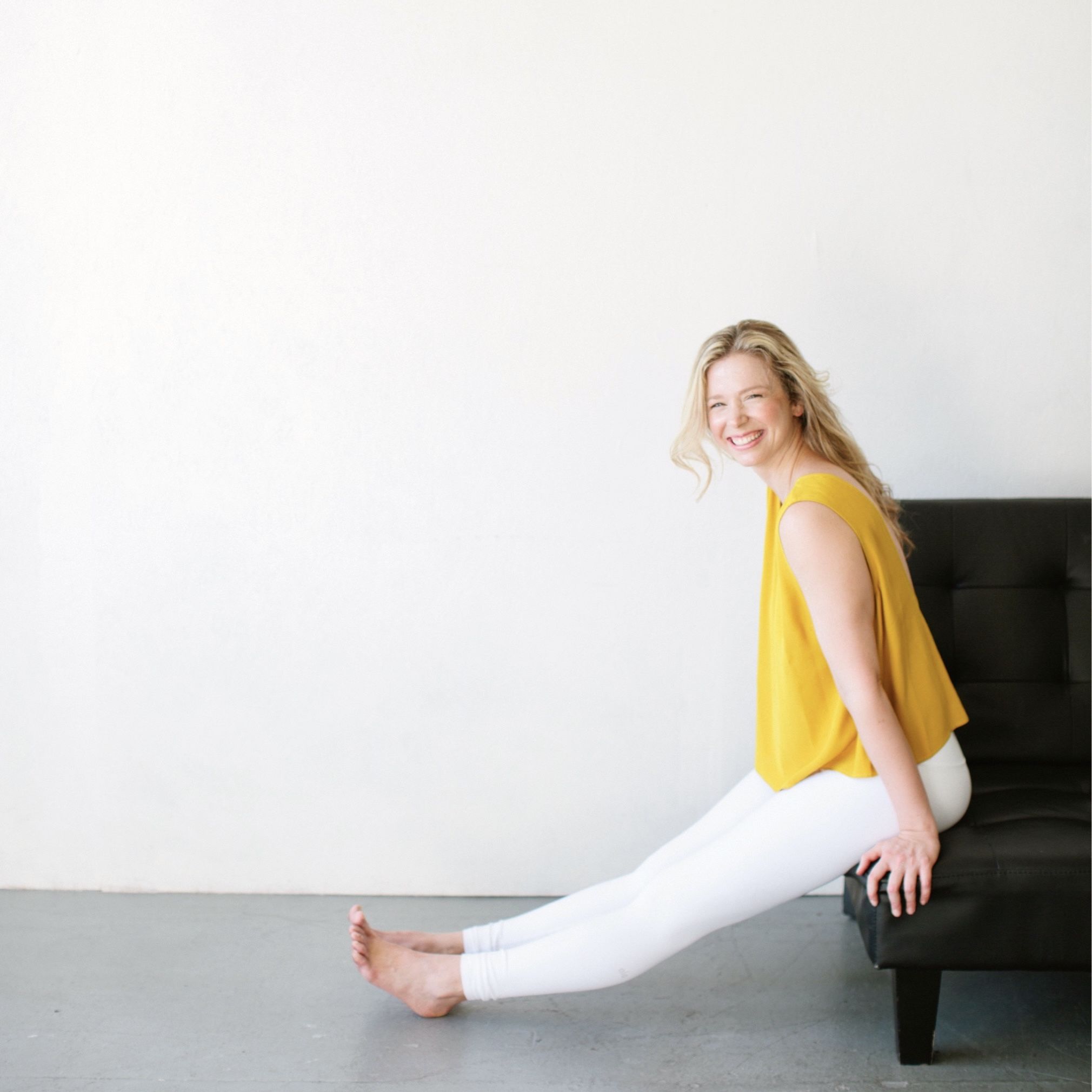 woman in yellow shirt and white pants sitting on black leather couch smiling at camera, white wall, grey floor