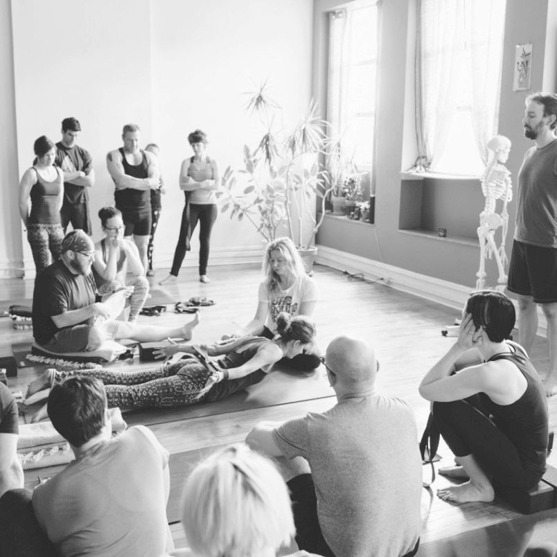 black and white photo of a yoga class, yoga teacher working with a student and other students observing, light streaming through the windows