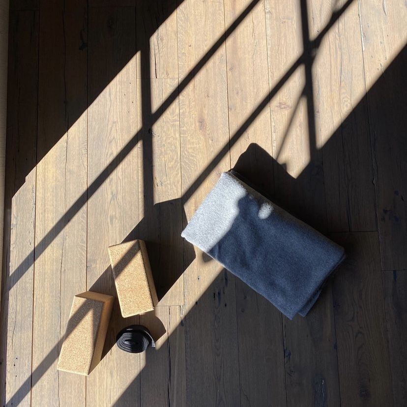 two yoga blocks, one blue yoga strap, and a grey folded yoga blanket on a wood floor with shadows of window panes on the floor