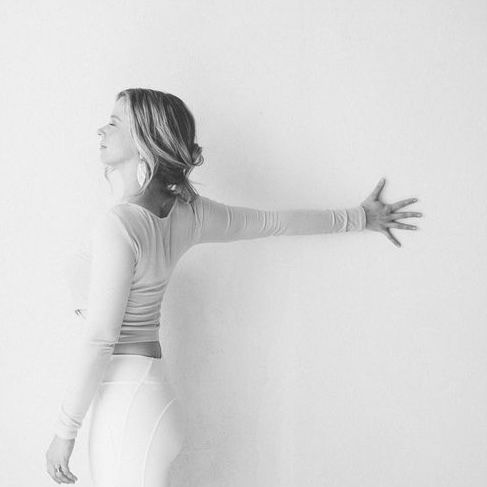 woman doing shoulder stretch at white wall wearing white pants and a long-sleeve top
