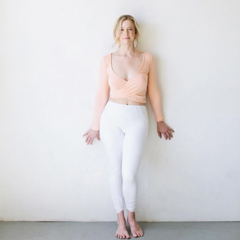 woman leaning against a white wall with a grey floor wearing white pants and a peach long-sleeve top