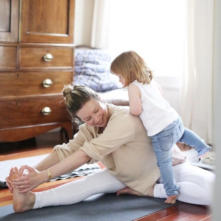 woman practicing yoga wearing white pants and neutral sweatshirt with toddler in jeans and white shirt climbing on her, wood armoire in the background