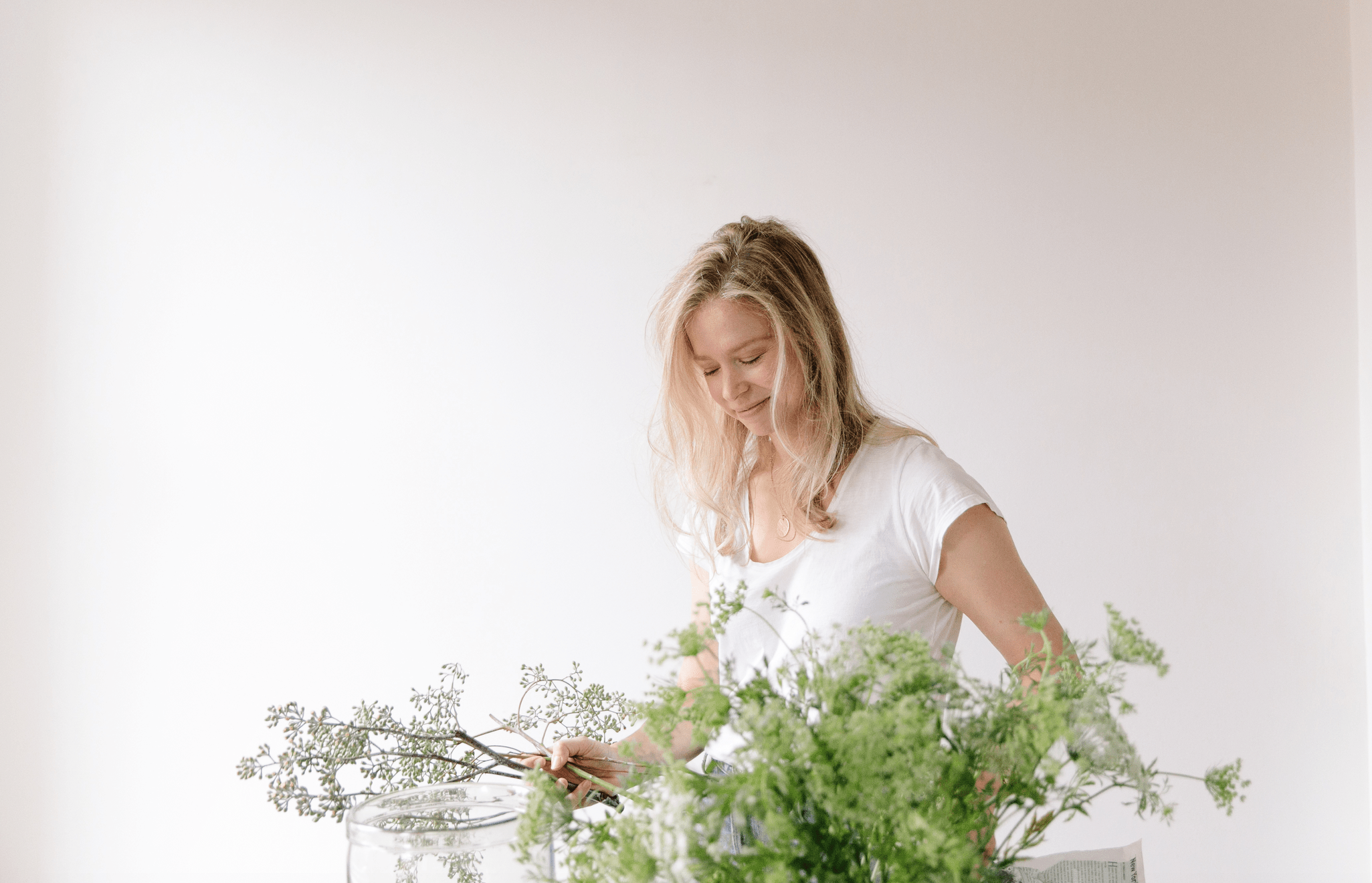 woman in white t-shirt against white wall arranging green flowers