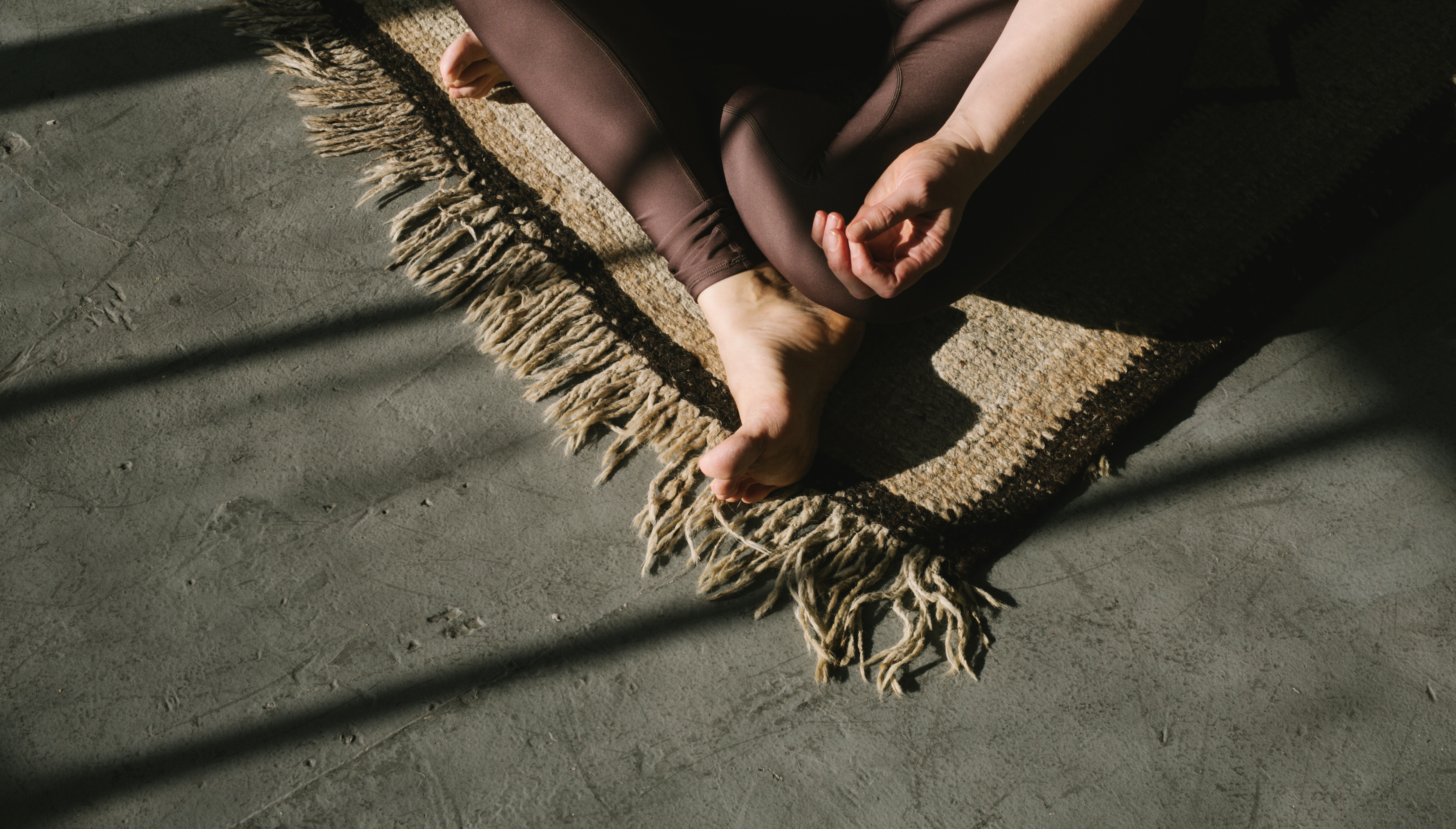 cross-legged seat wearing mauve-colored pants sitting on rug on top of concrete floor, light and shadows