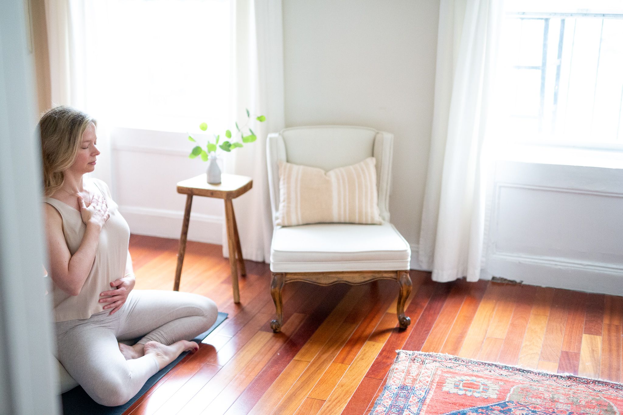 woman meditation wearing cream colors in room with chair and small stool with greenery in a vase