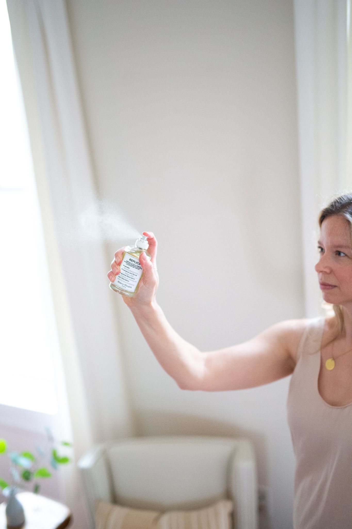 woman wearing beige in white room spraying perfume into the air