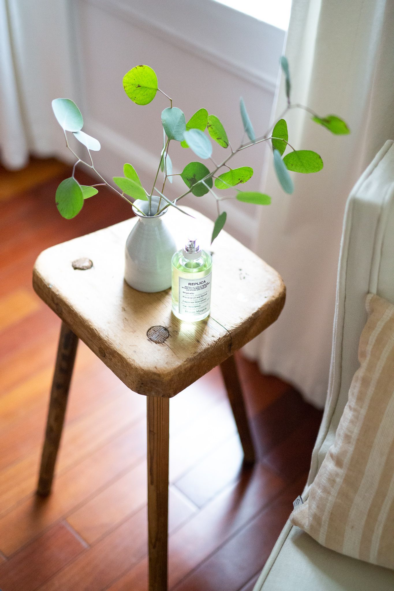 bottle of perfume sitting on wooden stool with vase of green leaves, chair and pillow in foreground