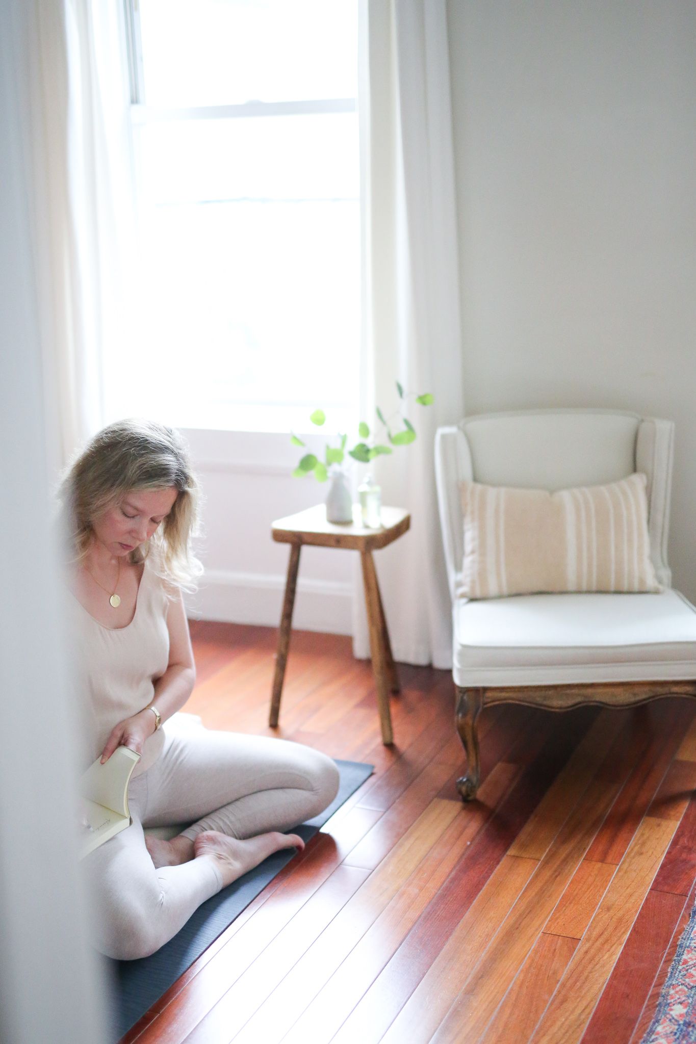 woman journaling wearing beige sitting in cross-legged seat in room with small chair, pillow, stool with flowers
