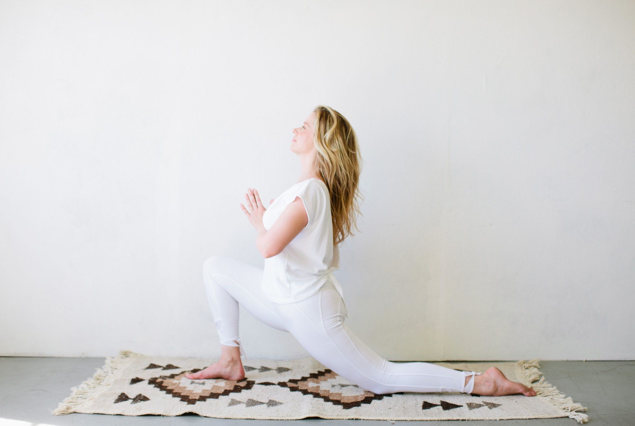 woman in lunge yoga pose wearing white practicing on patterned rug against white wall