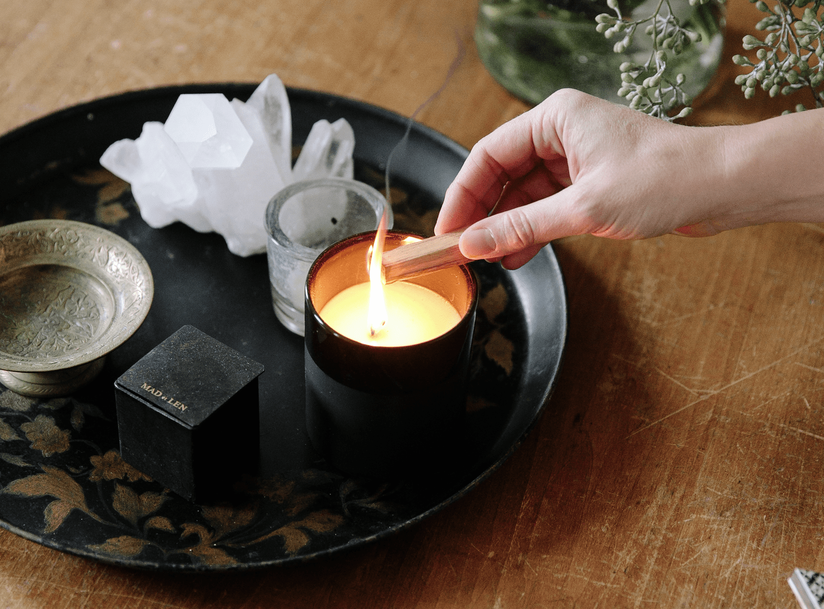 hand lighting incense, lit candle, crystal and brass bowl on black decorative tray resting on wooden table