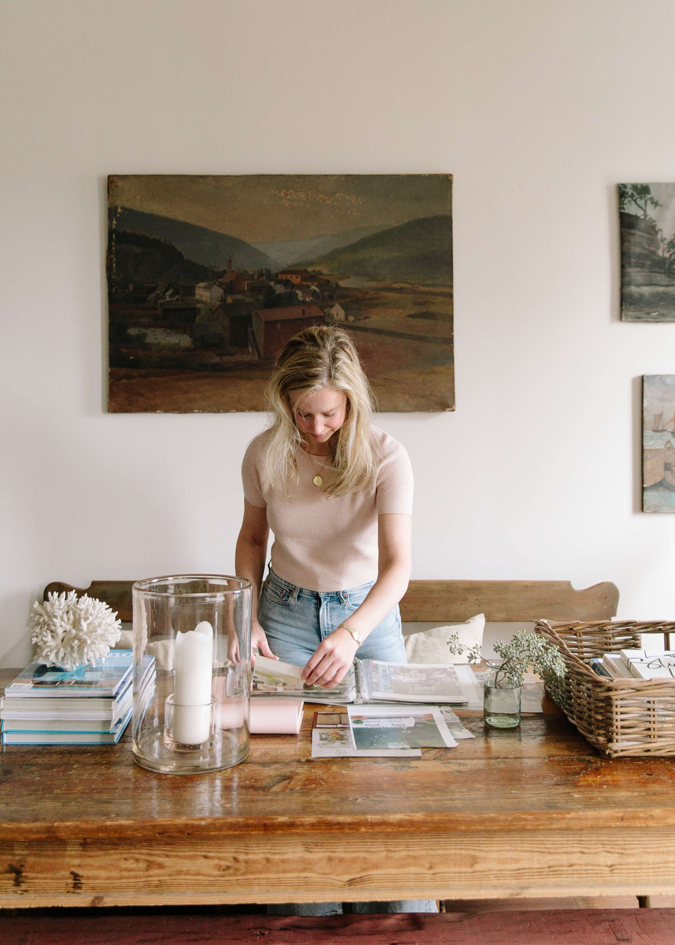woman standing behind wooden table, dark painting on the wall behind her, she's wearing blush short sleeve sweater and jeans, flipping through binders
