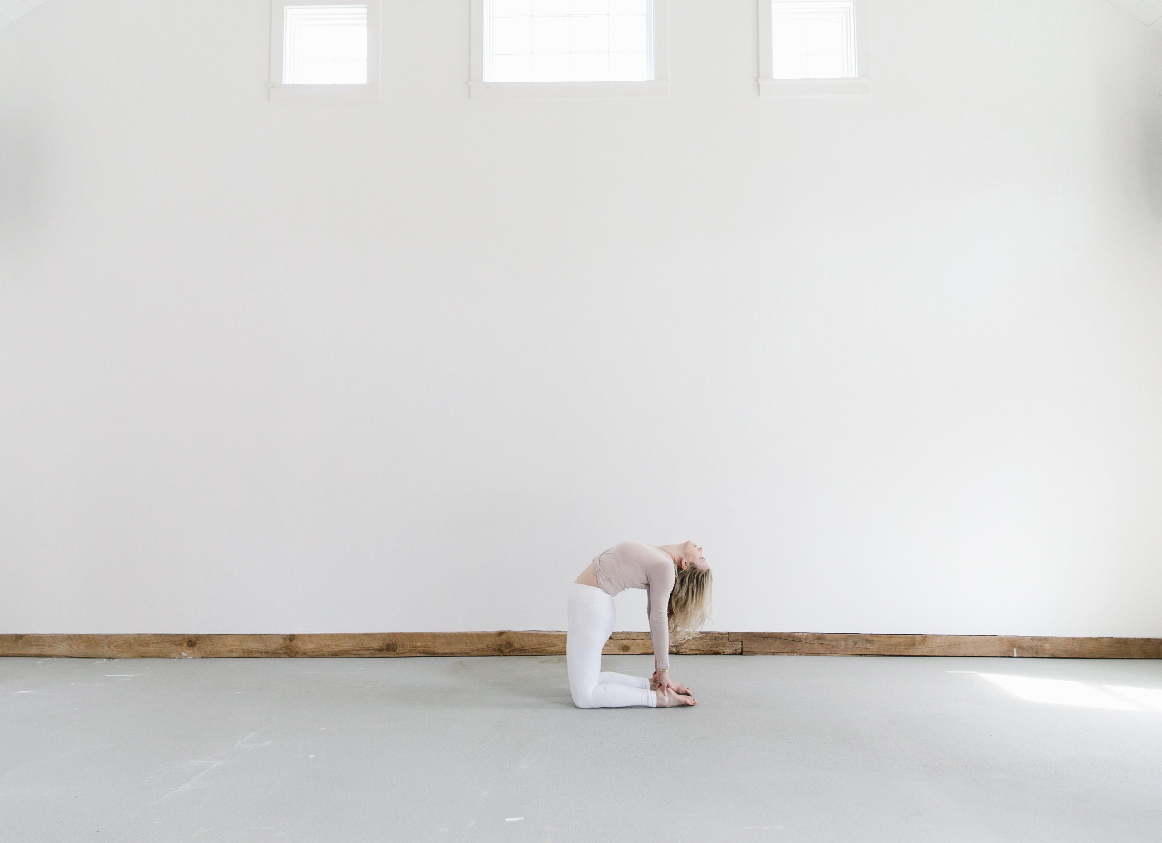 woman doing yoga pose in white pants and blush top against big white wall with three windows