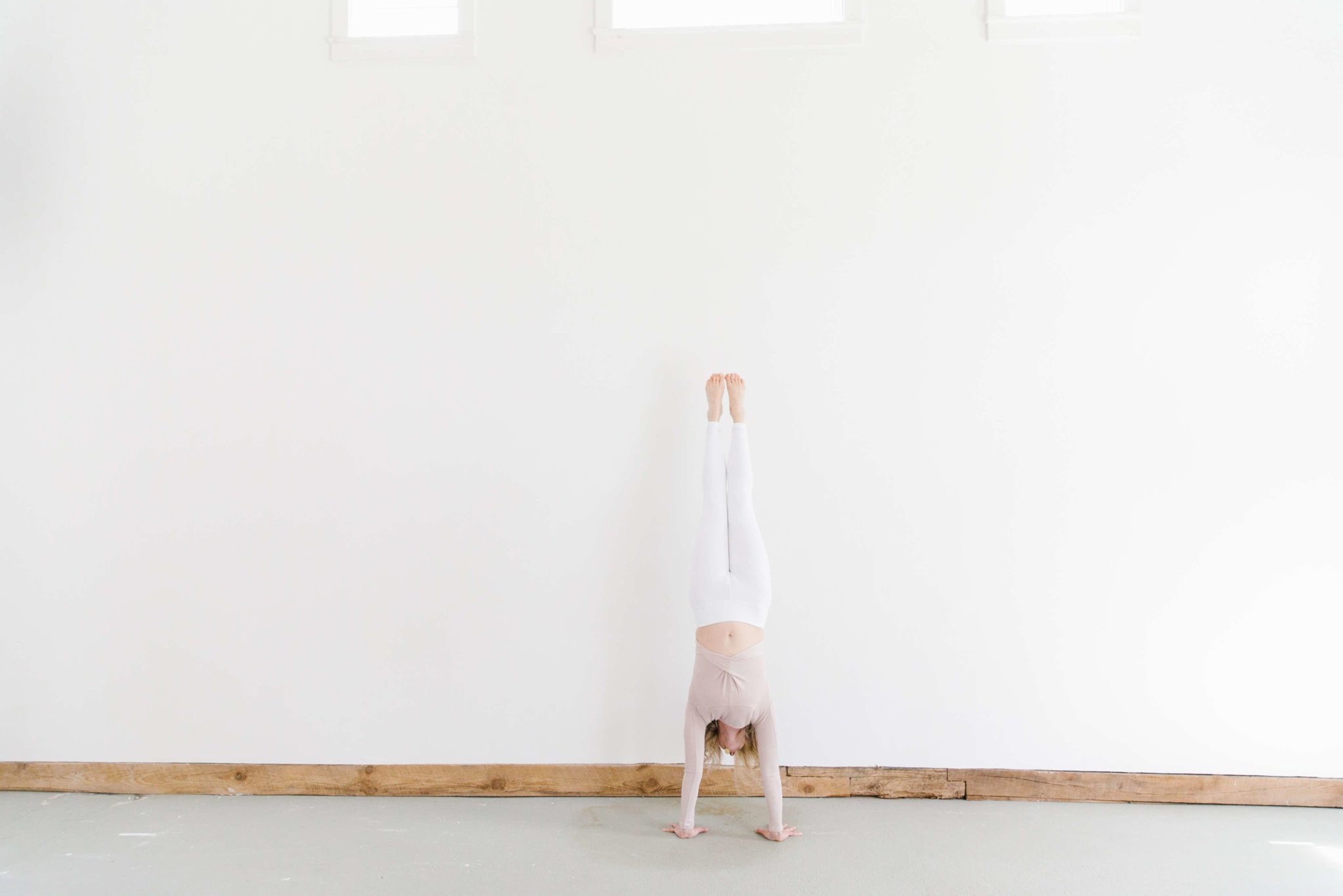 woman doing handstand in white pants and blush top against white wall