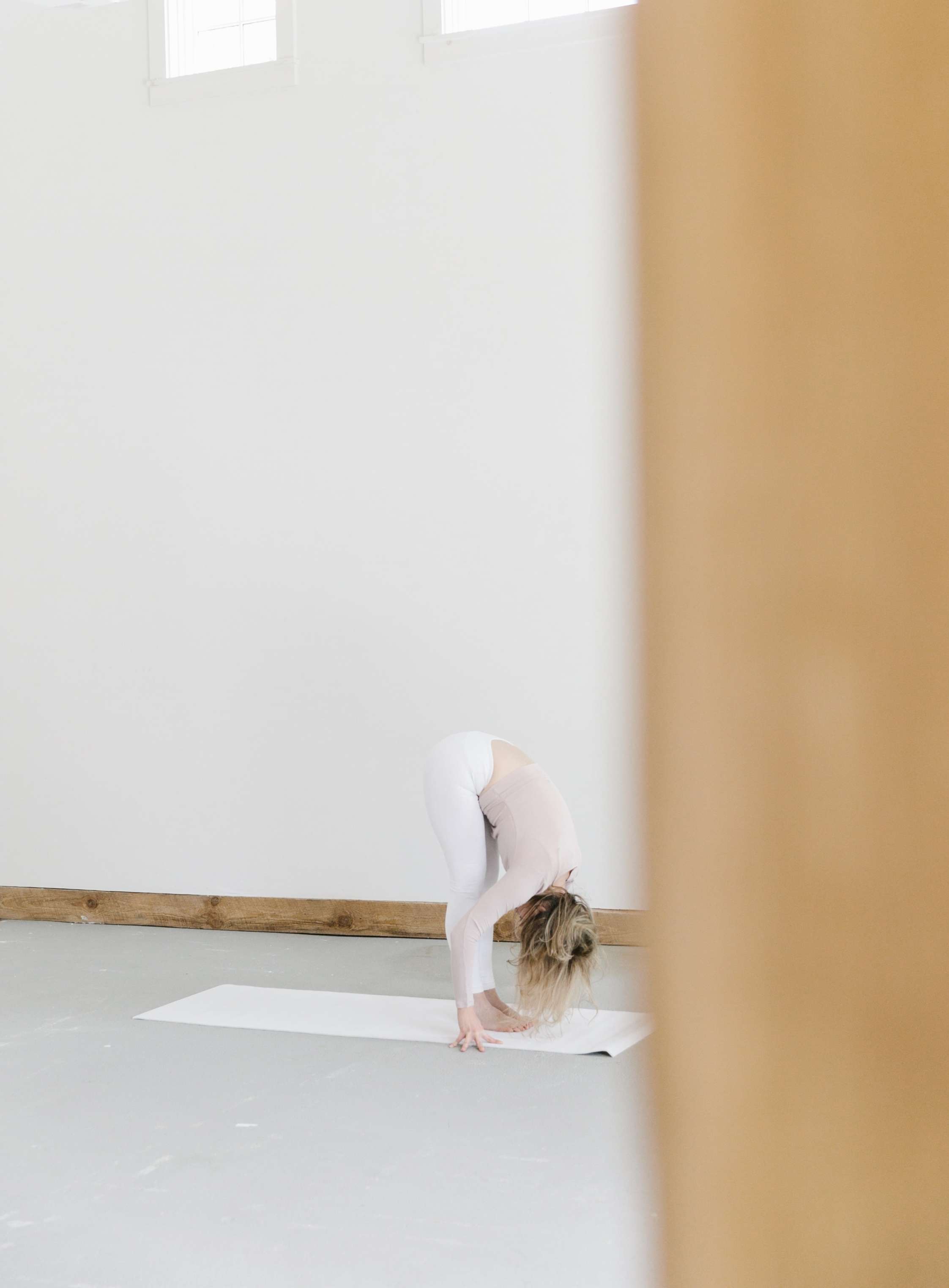 woman doing yoga forward bend behind brown pillar against white wall