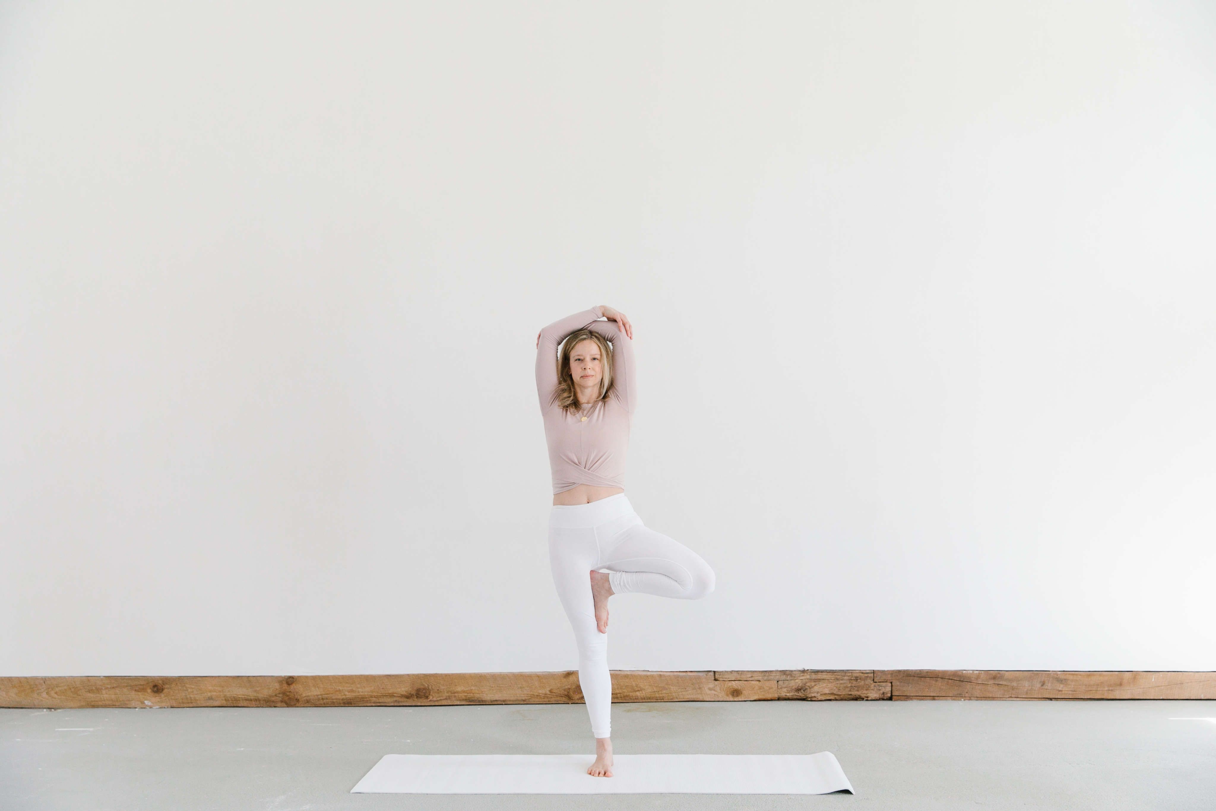 woman doing yoga pose named Tree in white pants and blush top against big white wall
