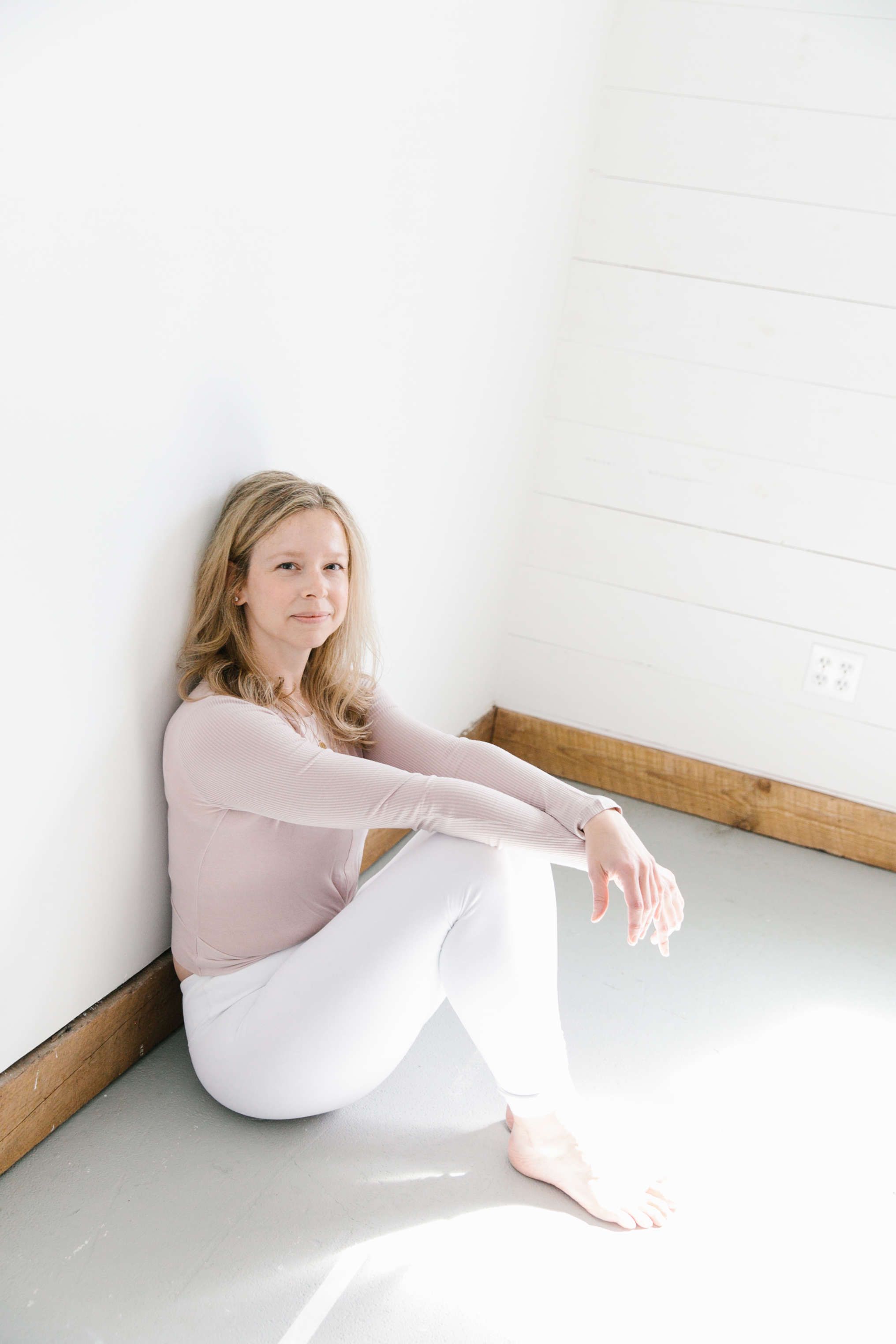 woman sitting on grey floor against white walls posing wearing white pants and a blush top
