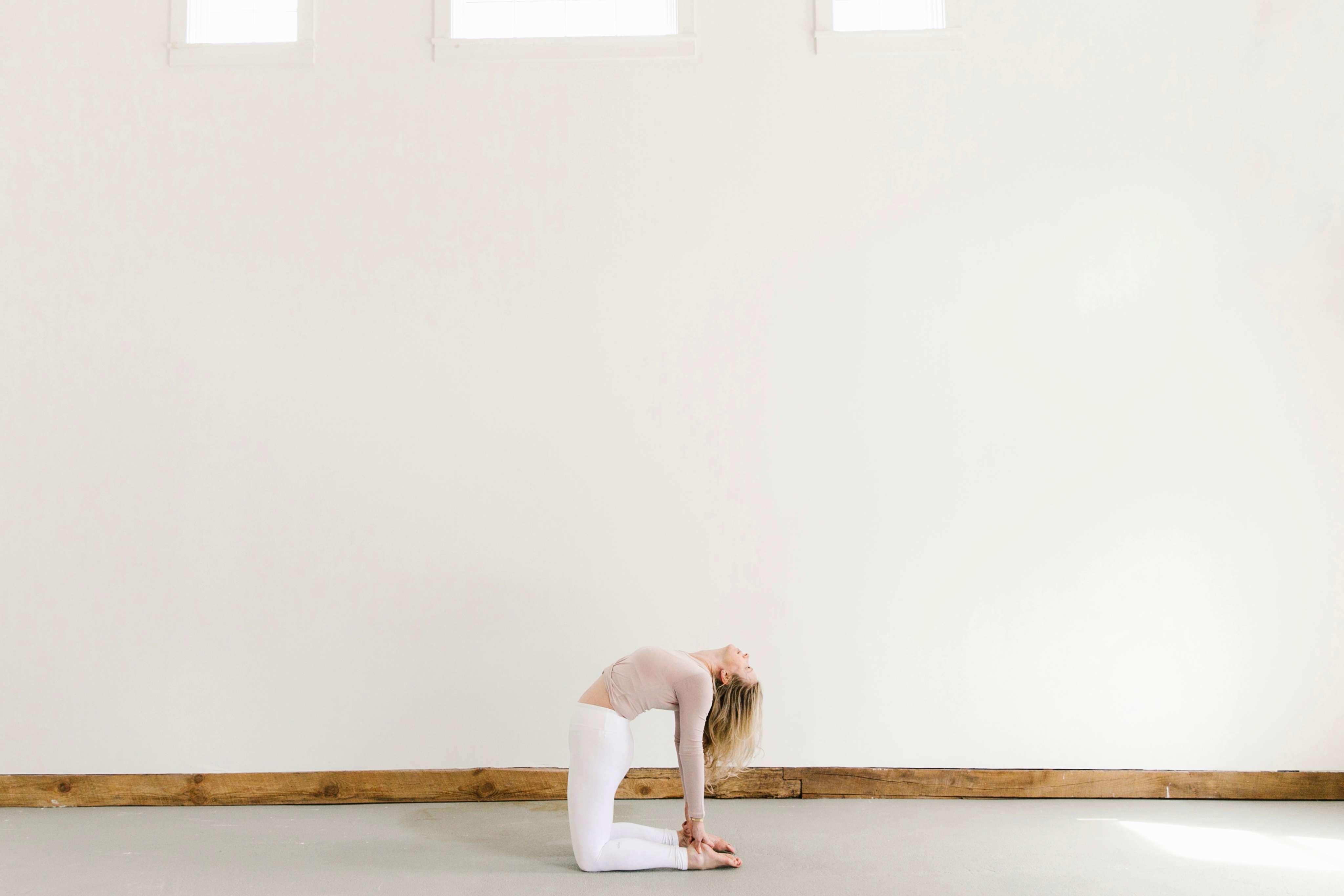 woman practicing yoga pose called Camel wearing white pants and blush top against big white wall with three windows above