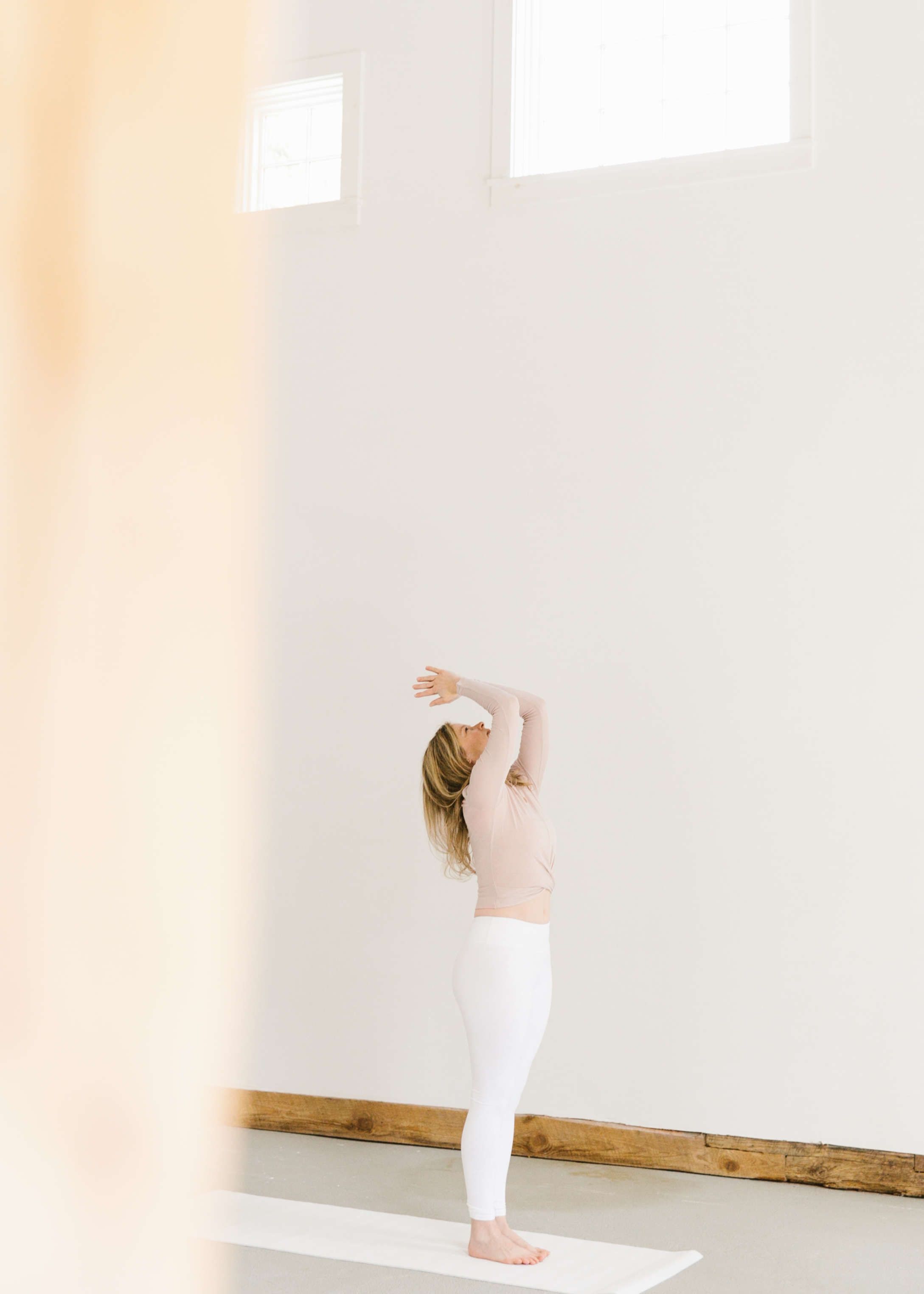 woman practicing yoga wearing white pants and blush top seen behind pillar against large white wall, two windows above