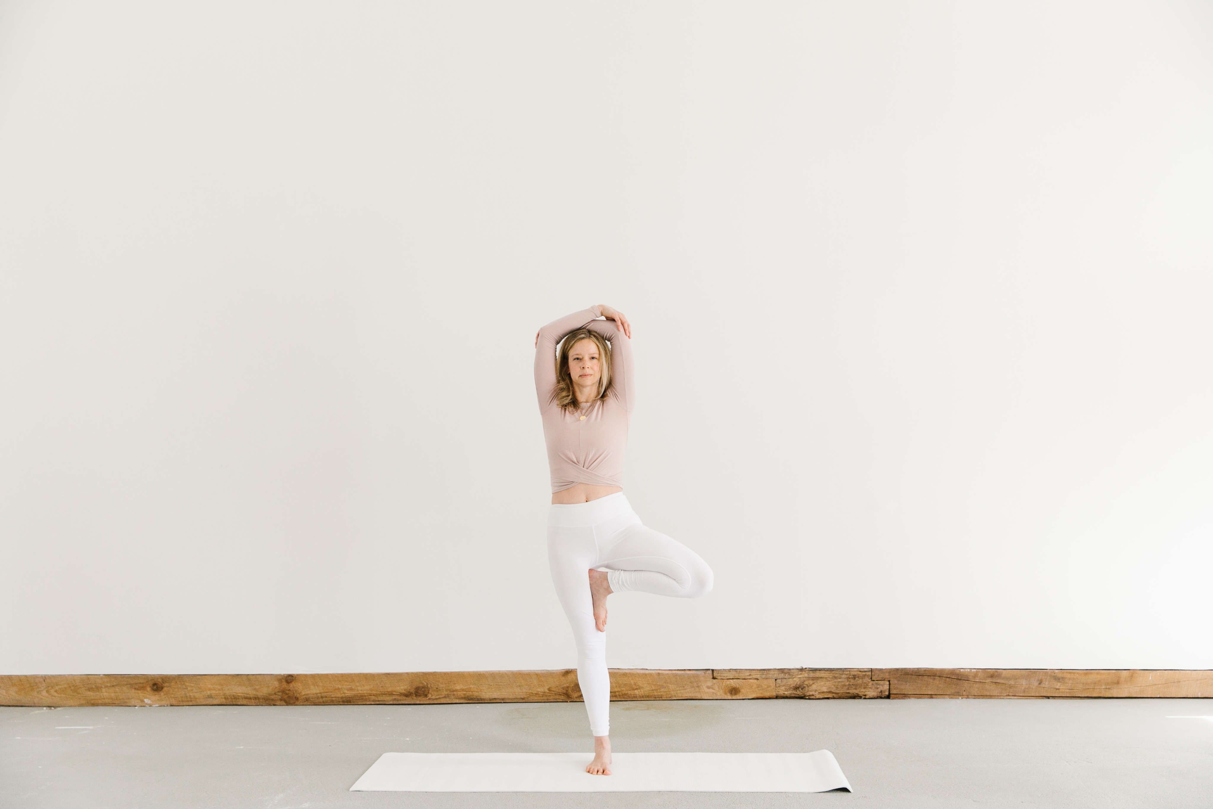 woman practicing yoga pose in white pants with blush top on white mat against white wall