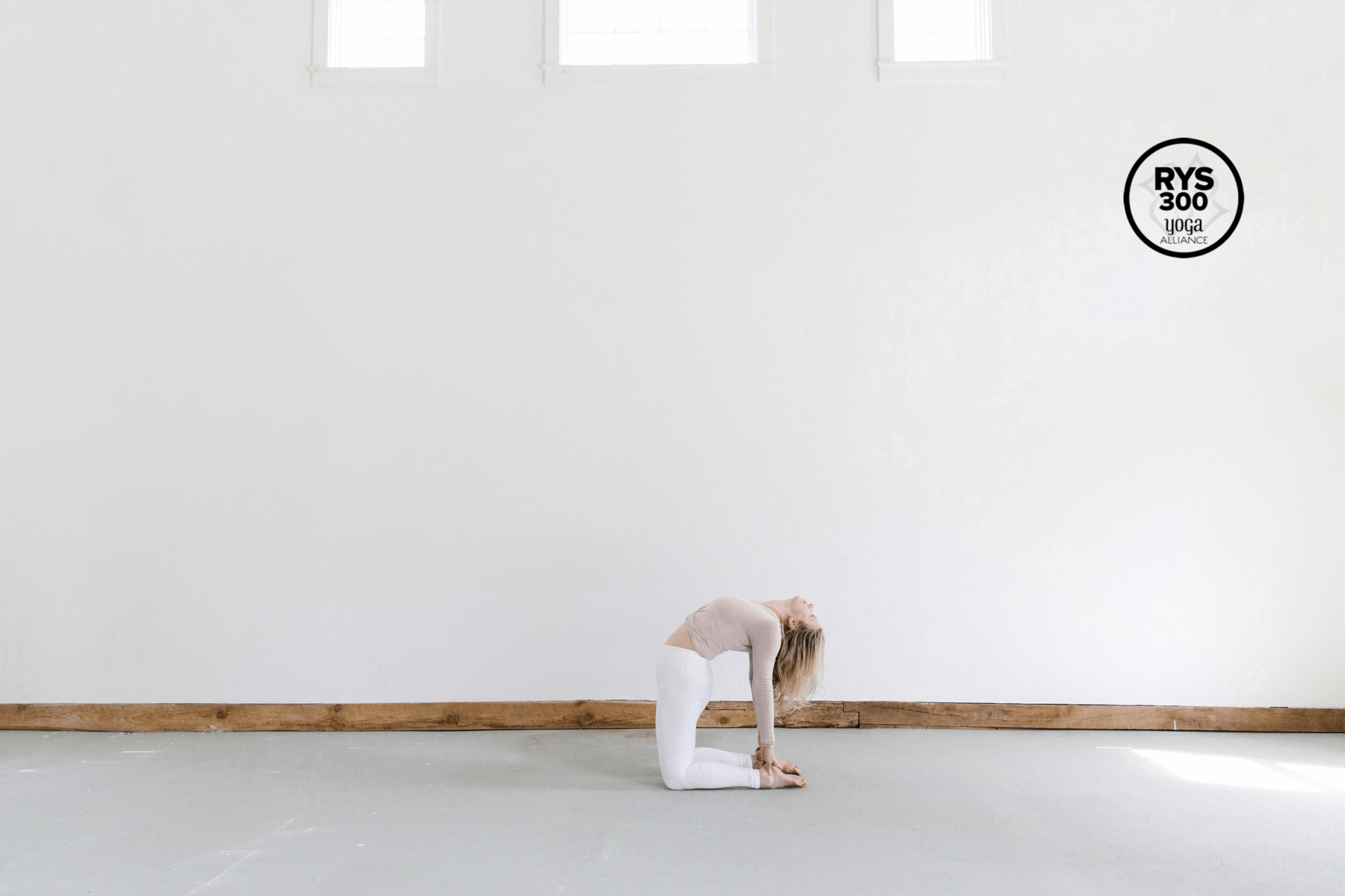 woman doing yoga pose in white pants and blush top against big white wall, logo reads RYS300