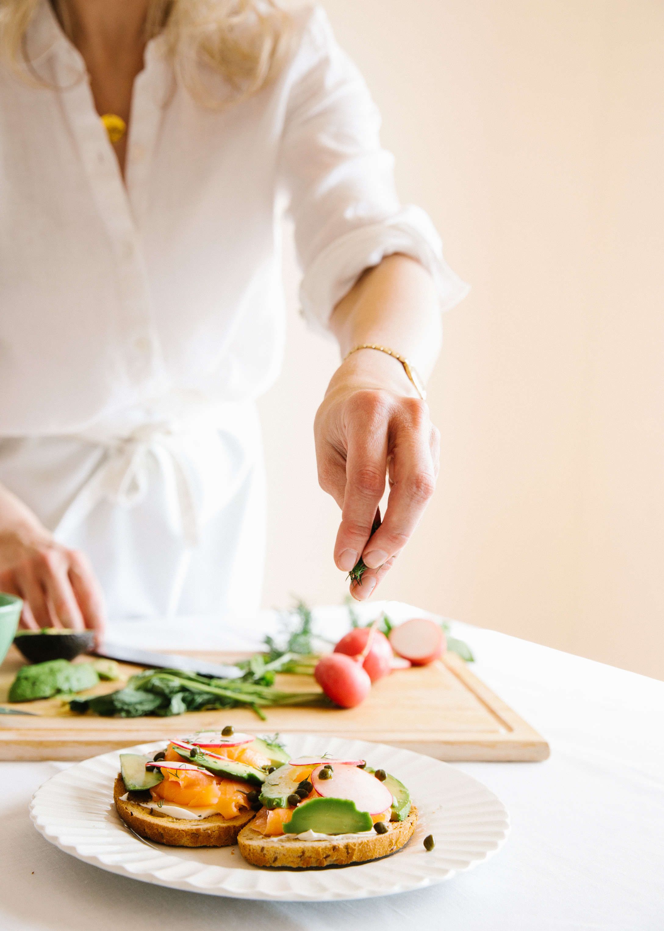 chef wearing white shirt and apron sprinkling capers onto a tartine of salmon, avocado, and radishes on white plate with cutting board in background