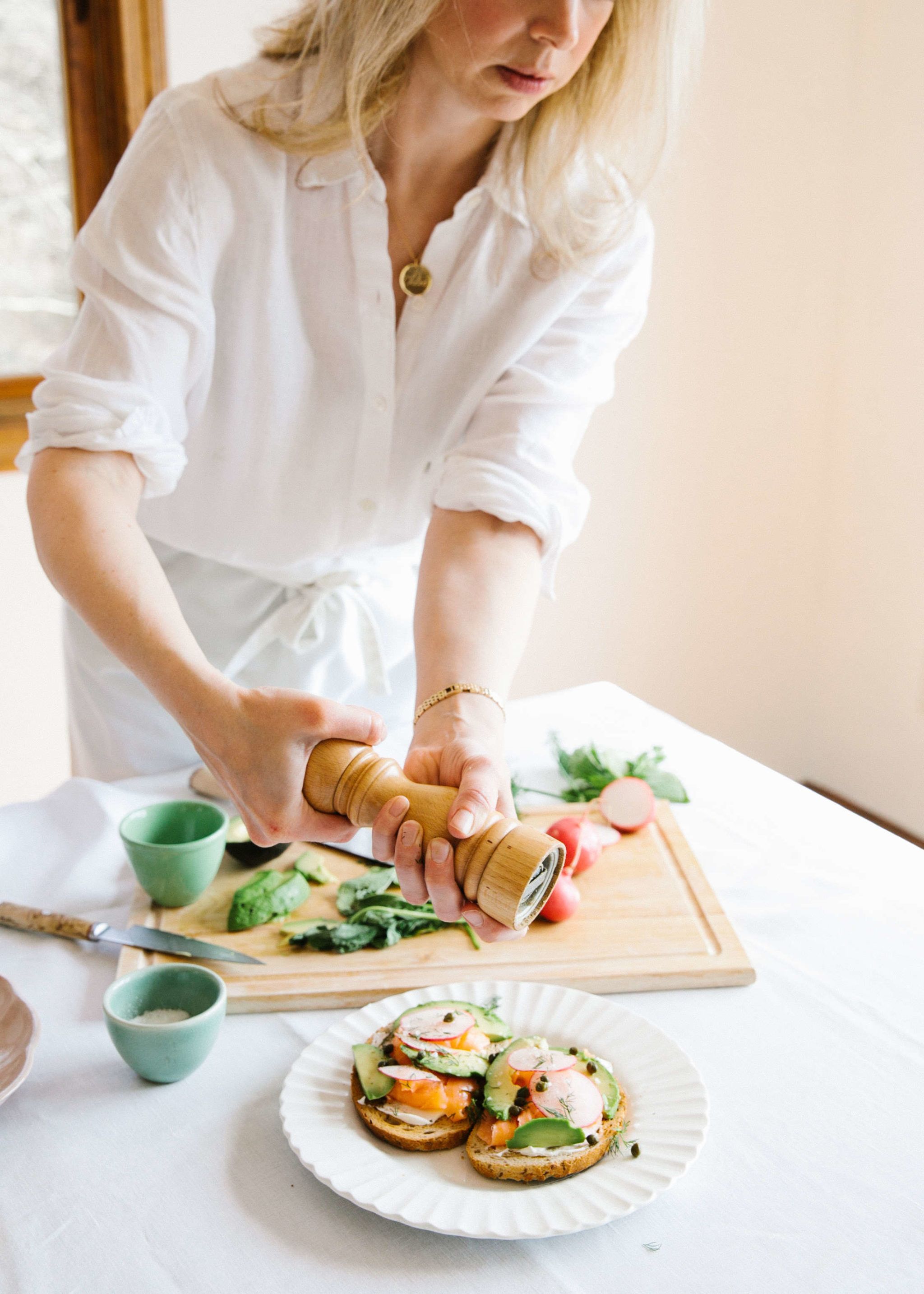 chef wearing white shirt and apron cracking pepper on top of tartine of salmon, avocados, and radishes on white plate with cutting board in background