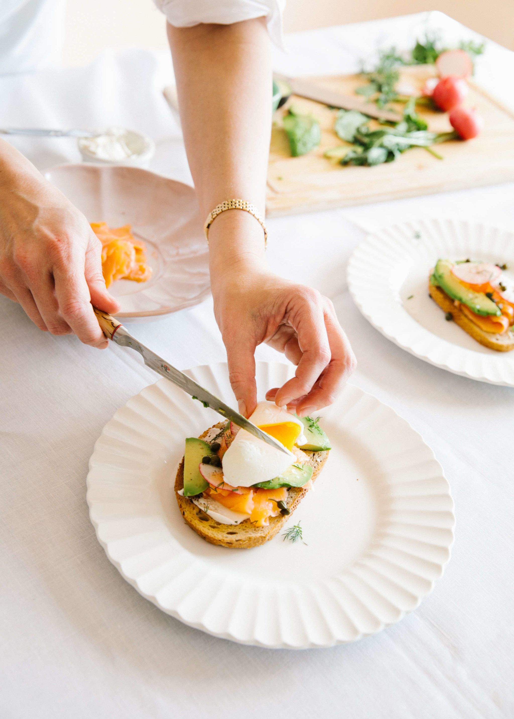 chef slicing into poached egg on top of tartine of salmon, avocado, and radishes on white plate with cutting board in background