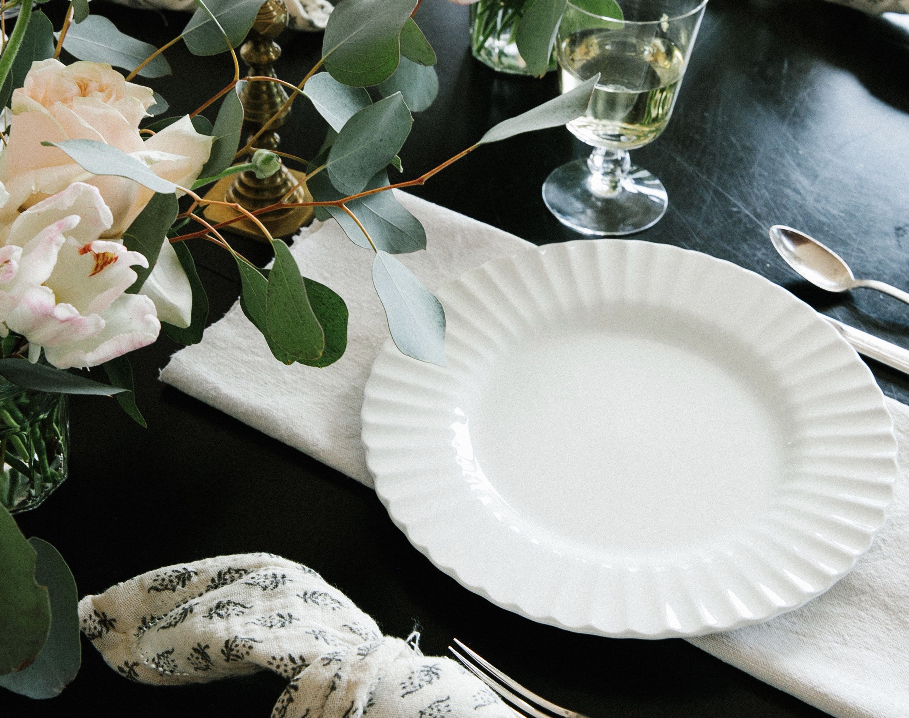 black table with table setting, white plate, patterned napkin, silverware, flowers, wine glass