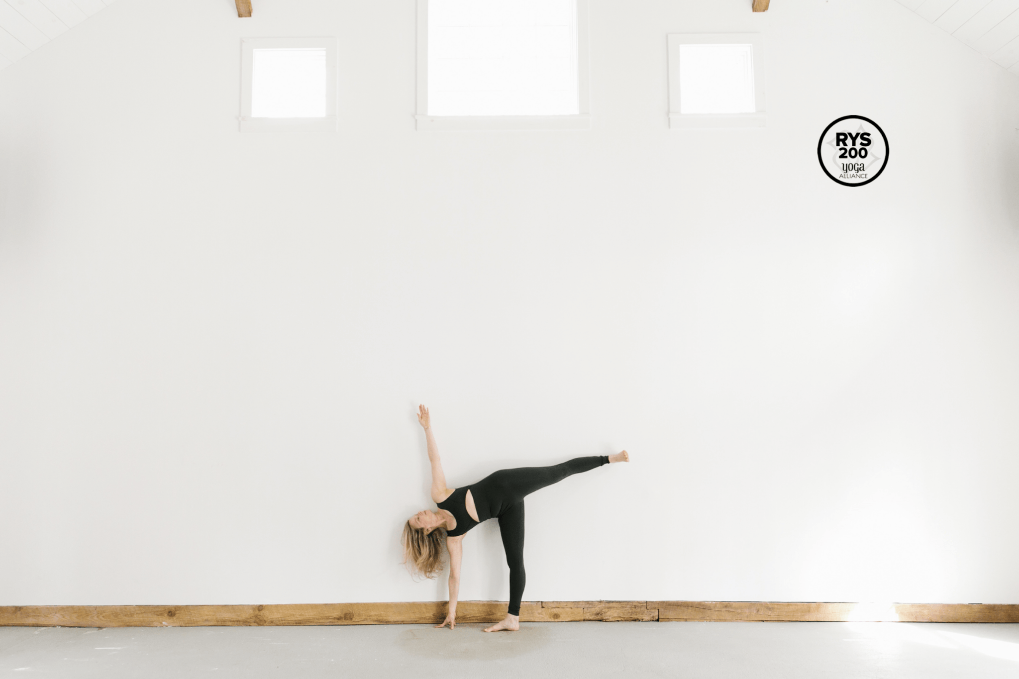 woman doing half moon pose in unitard against white wall with 3 windows