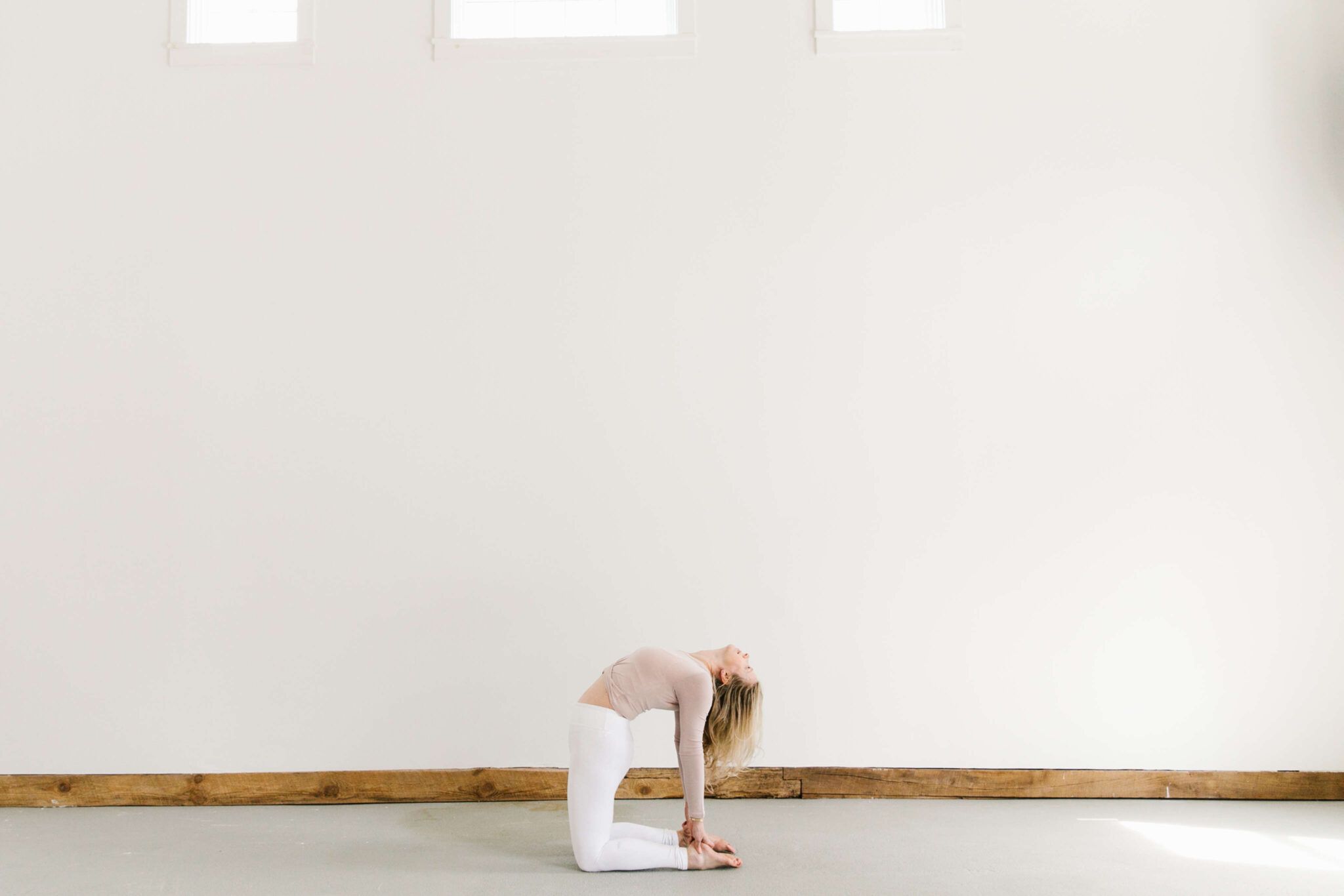 woman doing yoga pose backbend in white pants and rose colored top in large space with concrete floors, wood molding and white walls with three windows at top