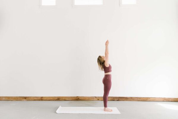 woman wearing maroon-colored yoga outfit doing a yoga pose standing with her arms up on a white mat against a white wall with three windows at the top of the frame