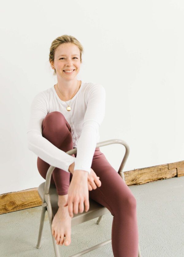 woman sitting in yoga chair wearing maroon yoga pants and white long-sleeve top with one foot on the chair and wrists crossed over chair back smiling at the camera