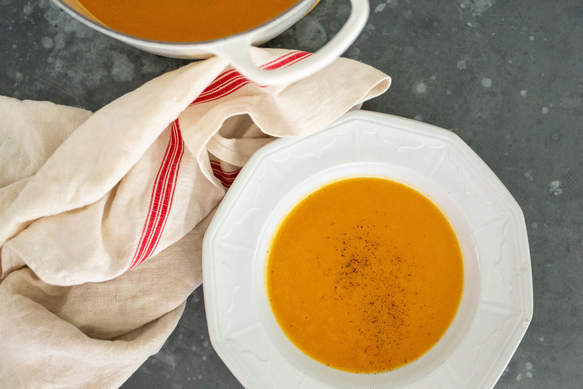 white bowl of orange butternut squash soup with fresh cracked pepper on gray counter with white tea towel that has red stripe and a nearby white pot of soup