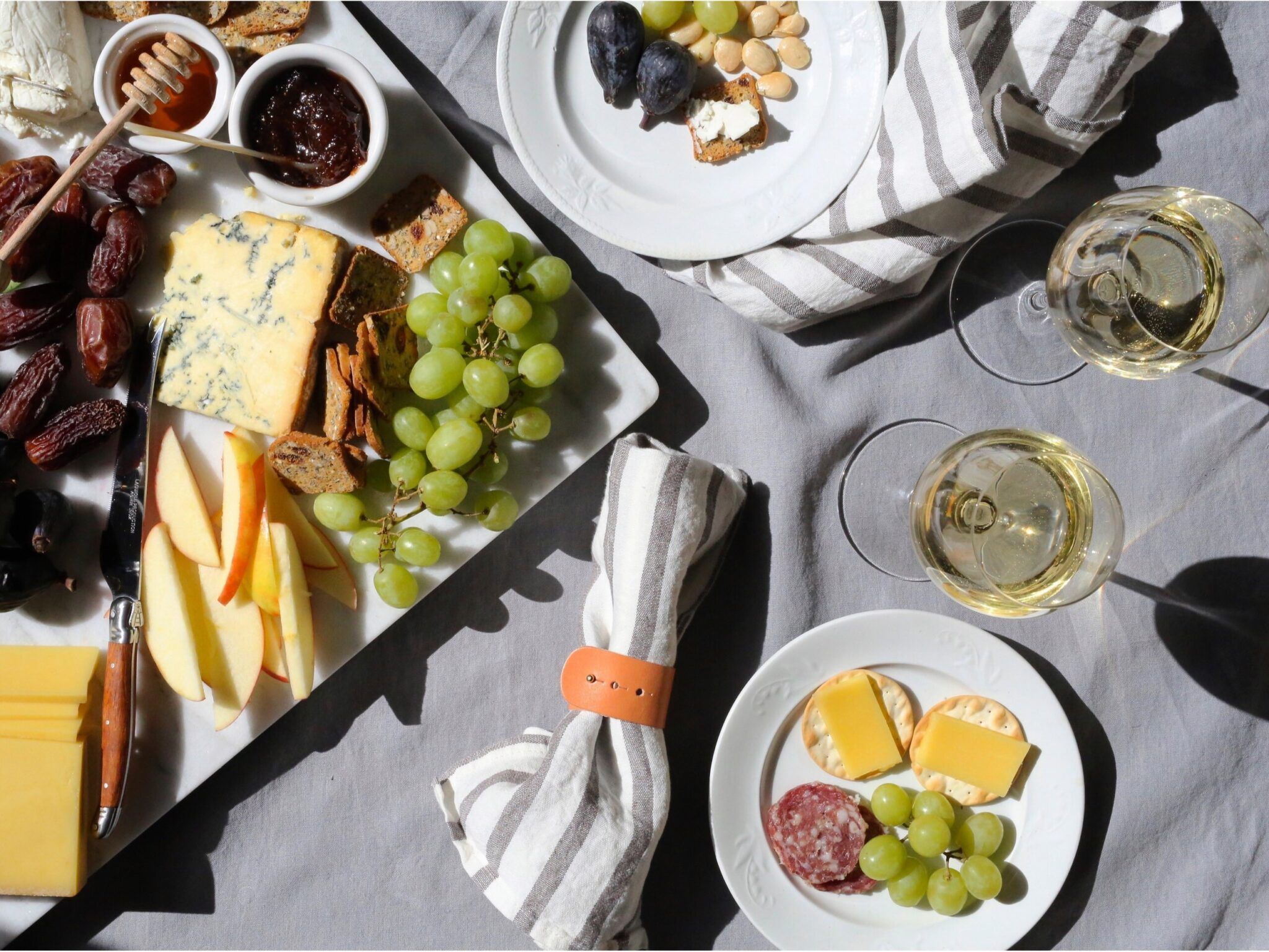 tablescape on blue tablecloth with cheese plate, striped blue napkins, 2 plates, 2 white wine glasses