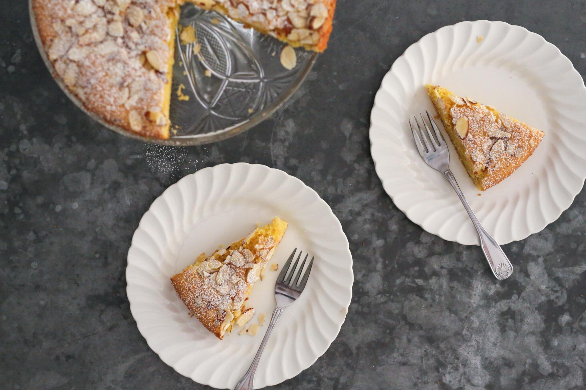 areal view of two plates with almond cake and forks with cake stand in upper lefthand corner
